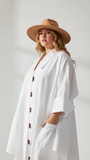 Woman wearing a white shirt dress  with brown buttons and a beige hat on a plain background
