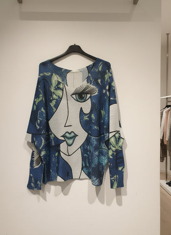 Blue sweater with artistic face design on a hanger against a beige wall