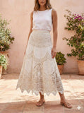 Woman wearing a white sleeveless top and lace skirt standing in front of a beige wall with plants.