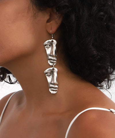 Close-up of a person wearing silver face-shaped earrings with a blurred background.